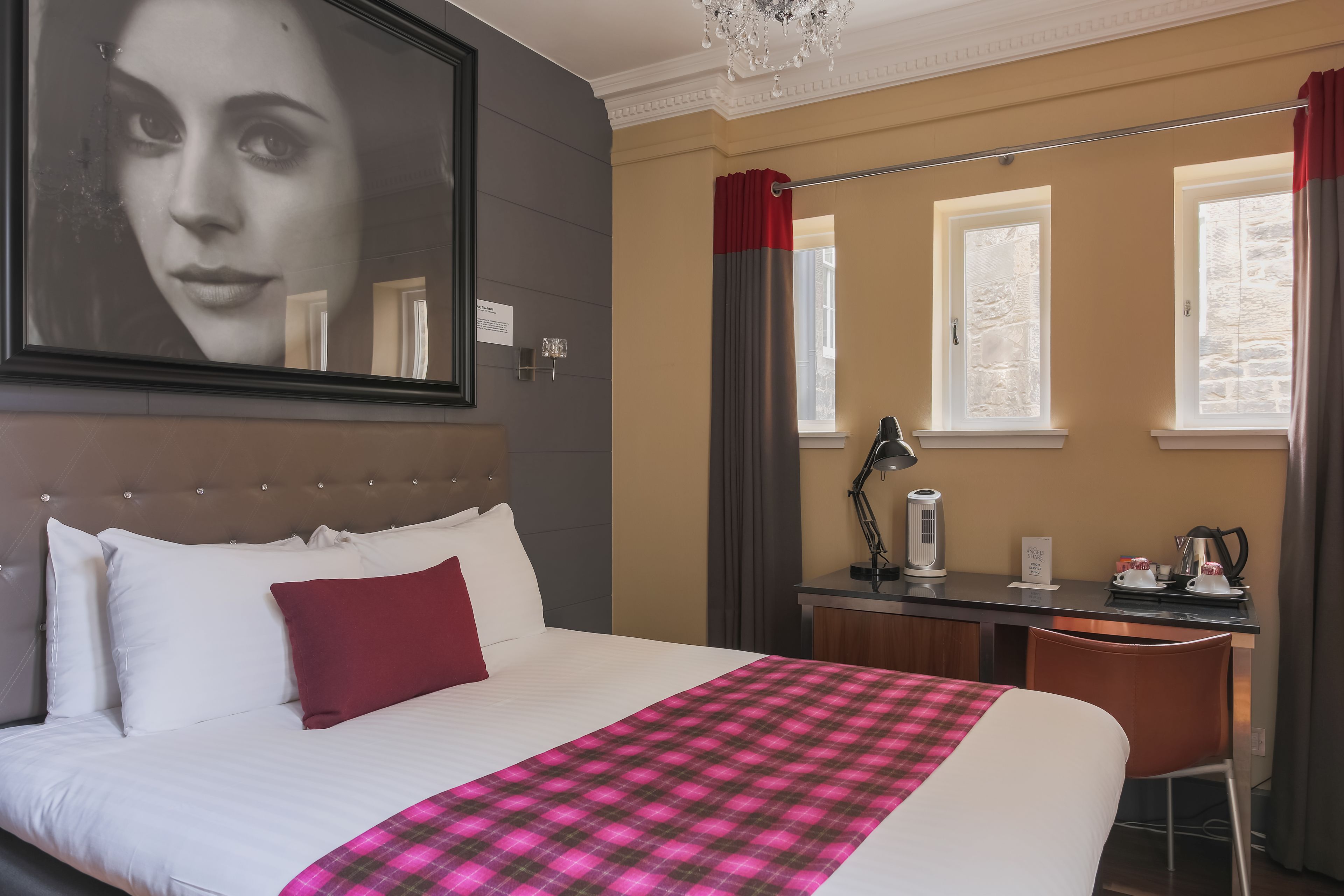 Habitación de hotel con una cama grande, un escritorio y una silla. Cuenta con una ventana con cortinas, una araña y un cuadro en la pared.
