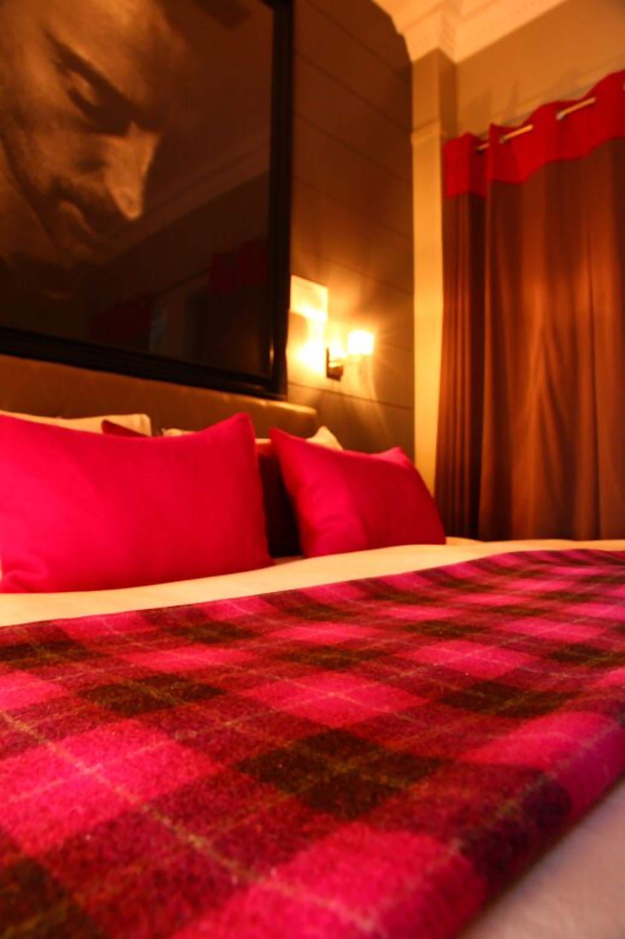 Habitación de hotel con una cama grande, almohadas rojas, una manta a cuadros, un televisor en la pared y una ventana con cortinas.