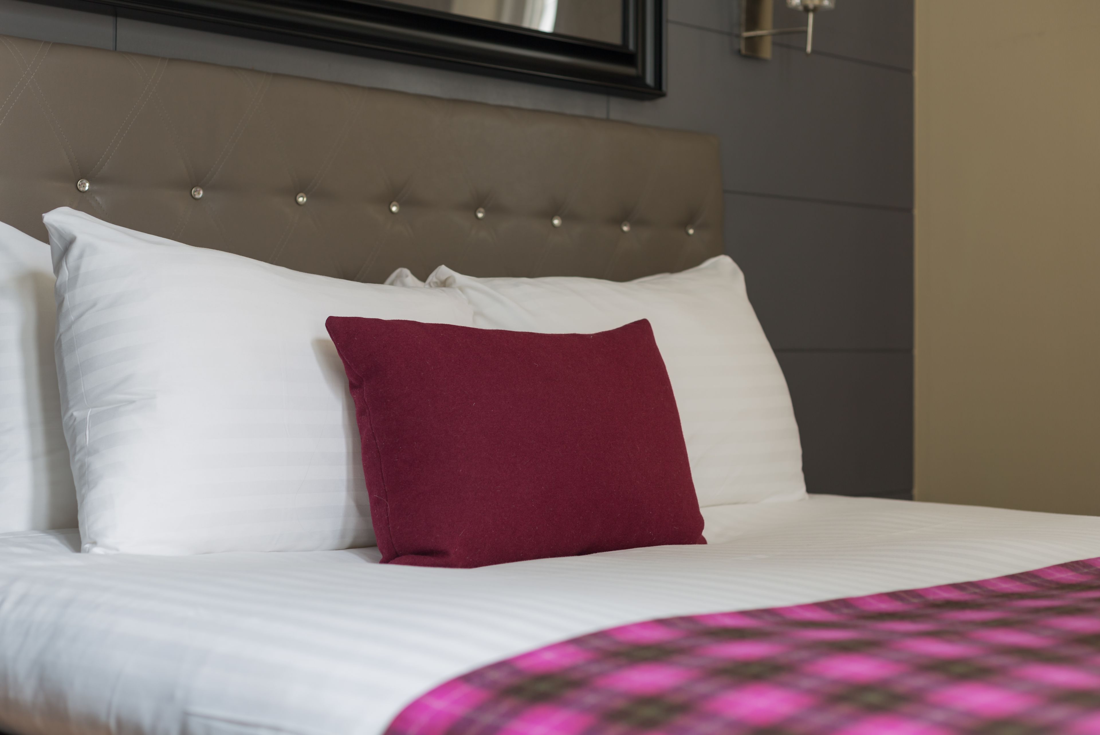 Una cama bien tendida con almohadas blancas y granate, un cabecero capitonado y una manta a cuadros rosa y negra.