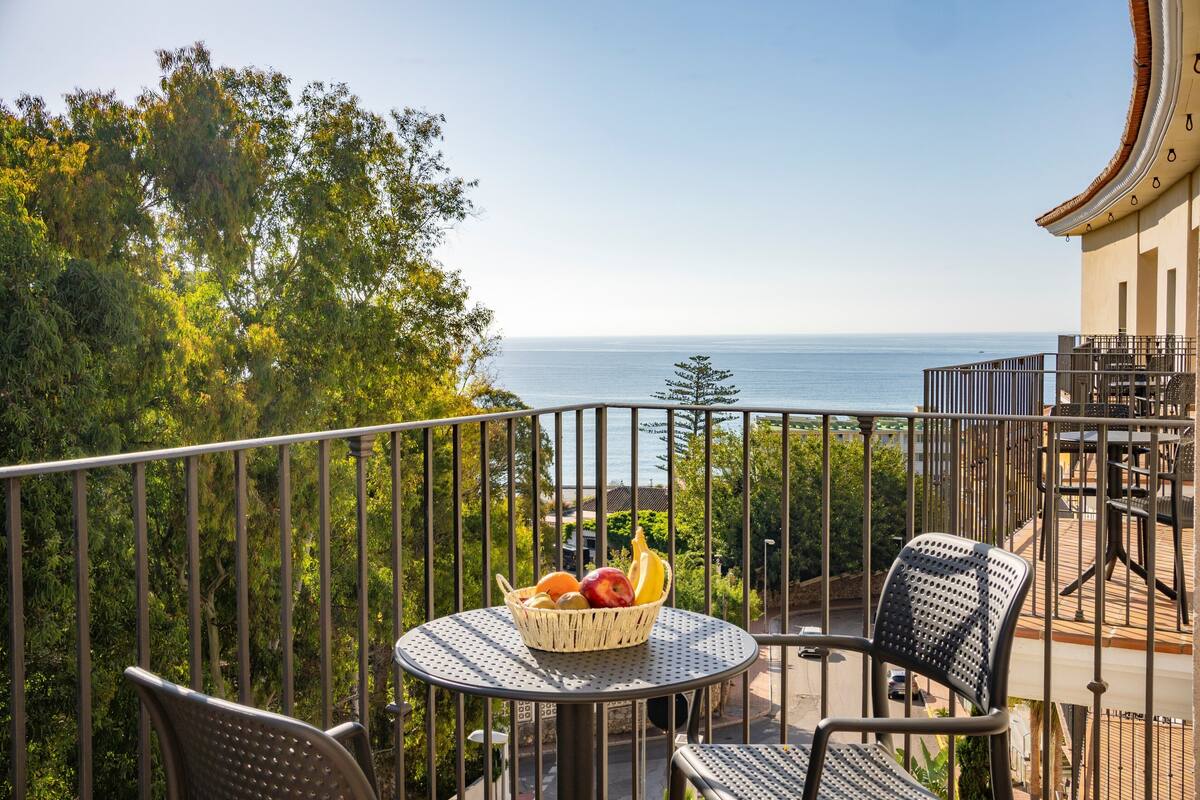 Un balcon avec une table sur laquelle est posé un panier de fruits, entouré de chaises, donnant sur une étendue d’eau et sur de la verdure.