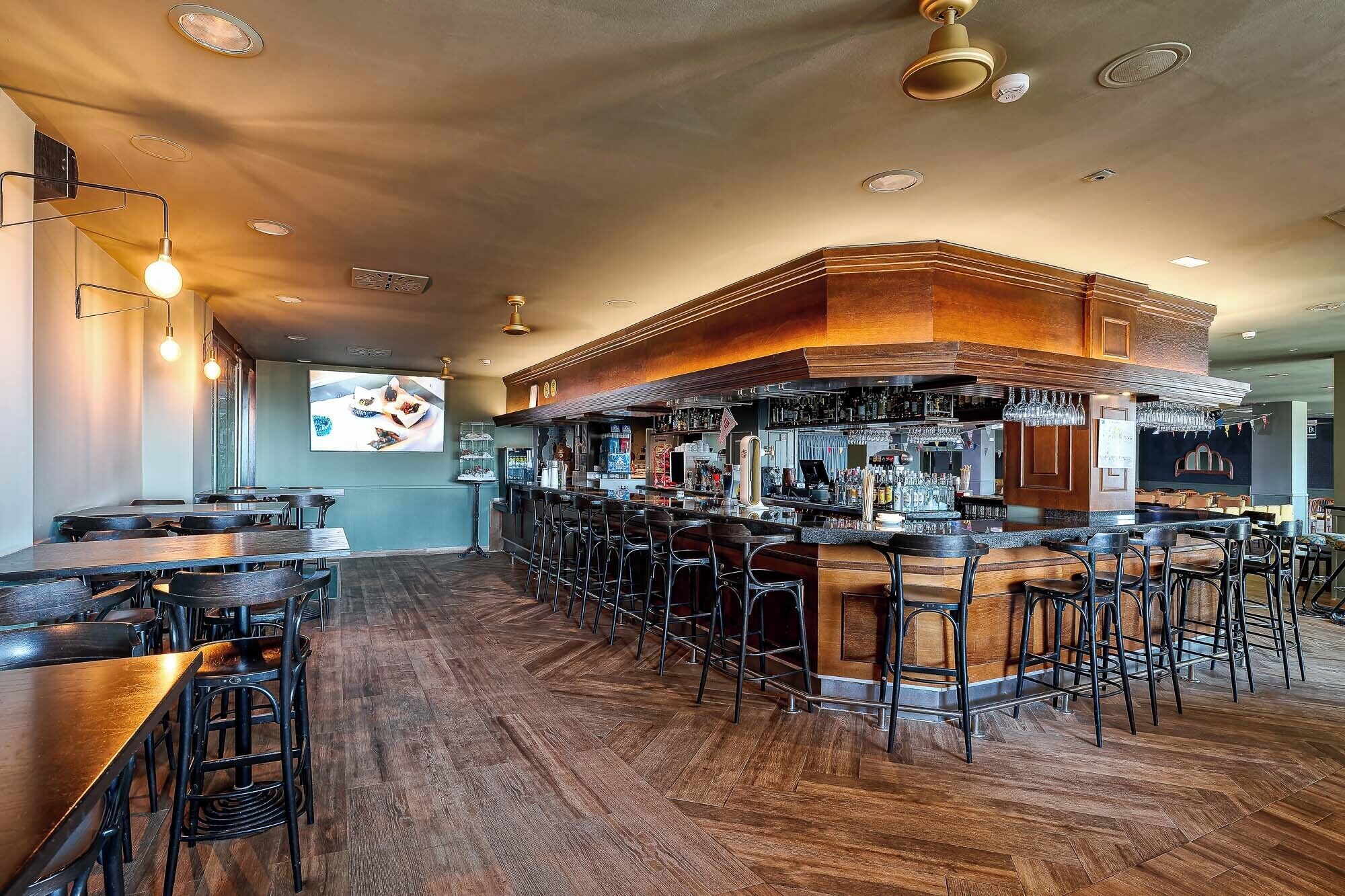 A bar area with wooden bar counters, multiple stools, and a large screen displaying food.