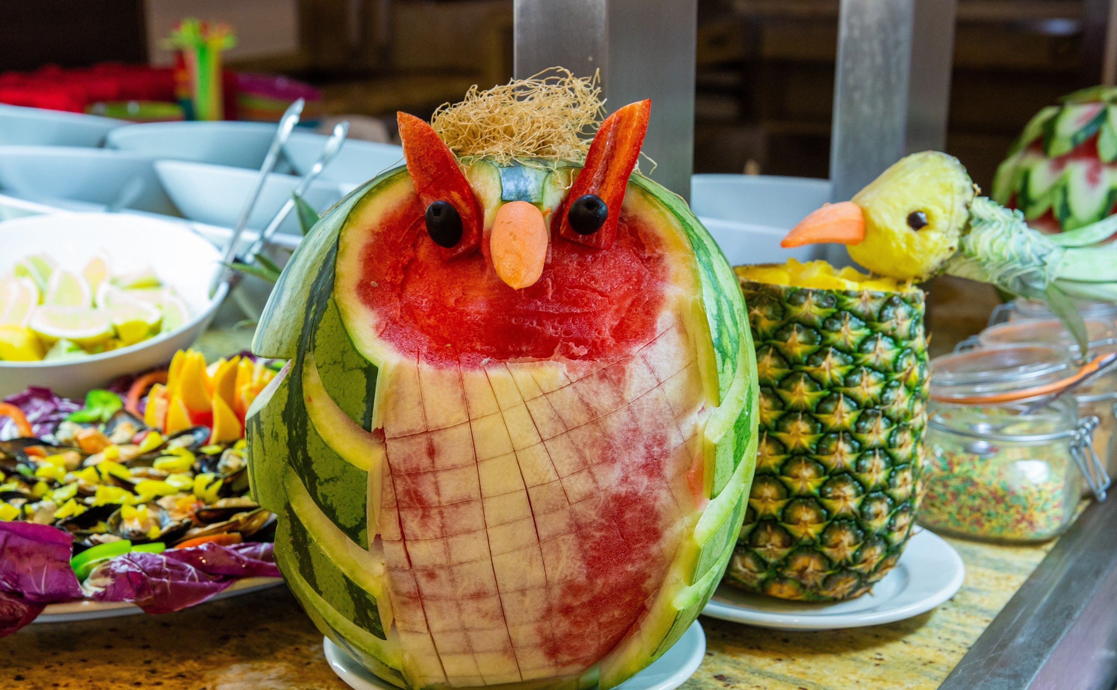 Una talla de fruta con forma de búho, con rostro y cuerpo detallados, rodeada de otras esculturas de fruta y una piña.