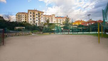 A sandy sports court with a netted fence, surrounded by greenery and buildings in the background.