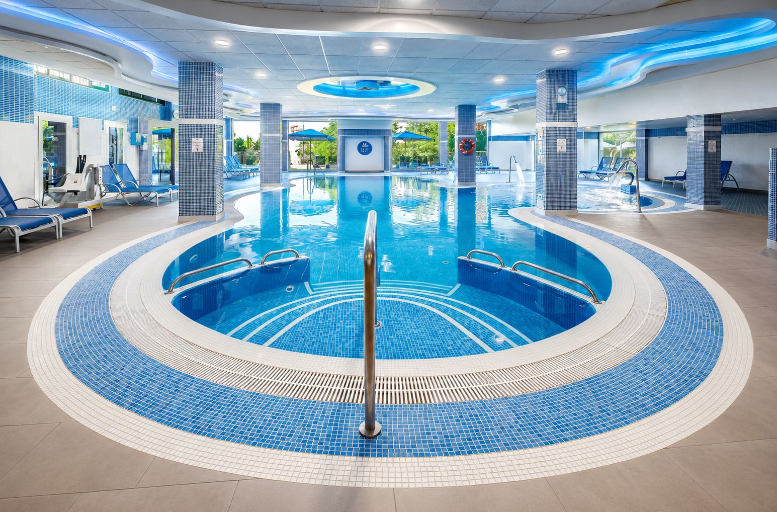 A circular indoor swimming pool with a central diving platform, surrounded by lounge chairs and a tiled floor.
