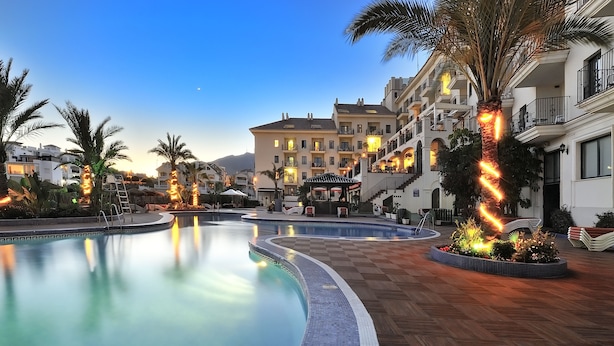Zona de piscina con borde curvo, palmeras y un edificio con balcones al fondo.