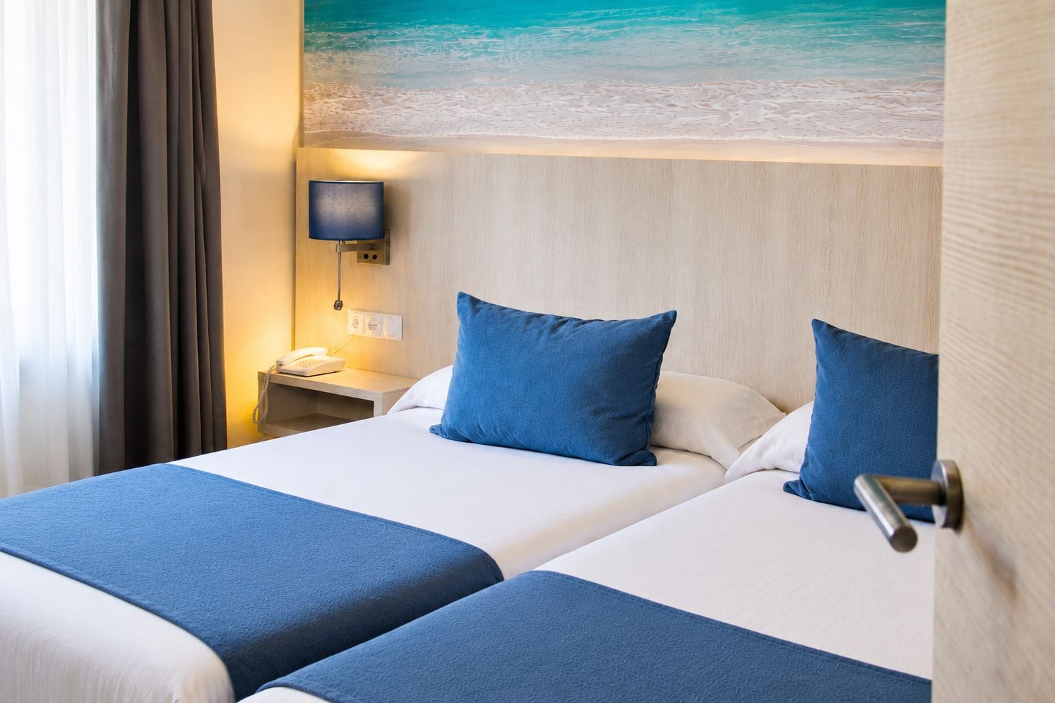 A hotel room with two beds, a nightstand, a lamp, and a wall-mounted phone.