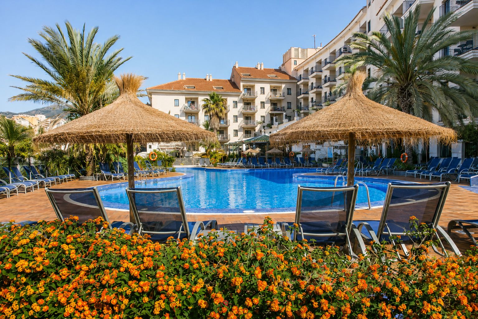 Une piscine entourée de chaises longues et dotée de parasols en chaume, le tout bordé d’un parterre de fleurs.