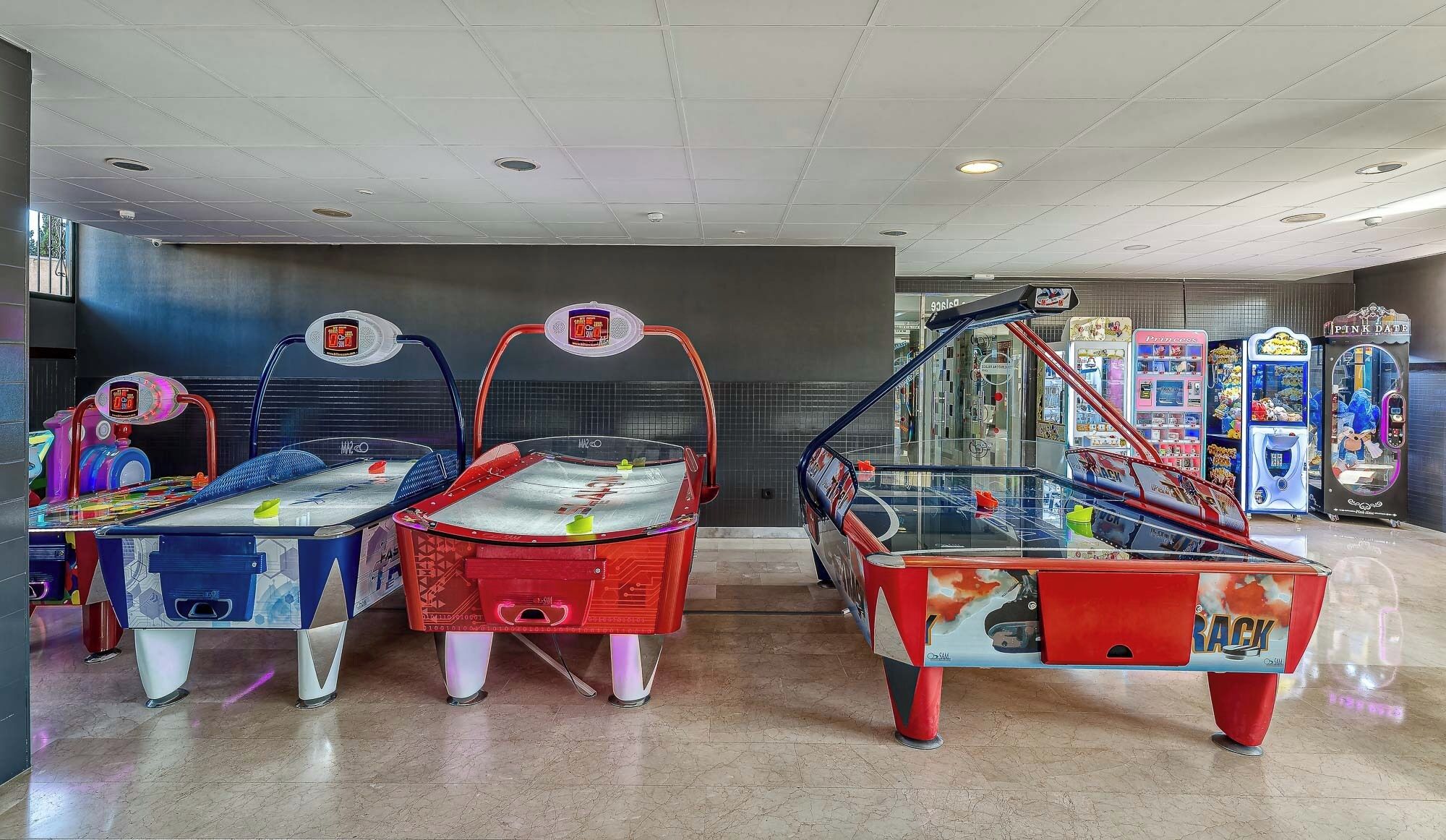 A row of arcade machines with a digital scoreboard, a glass display case, and various colorful vending machines in the background.