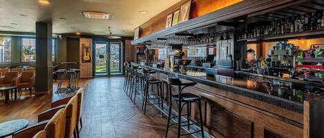 A bar area with wooden flooring, bar stools, and a variety of bottles and glasses on shelves.