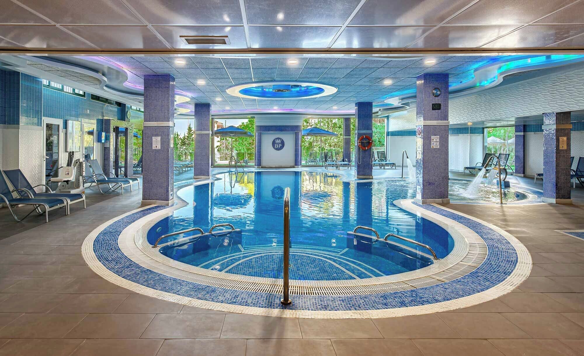 A large indoor swimming pool with a central circular hot tub, surrounded by lounge chairs and a tiled floor.