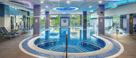 A large indoor swimming pool with a central circular hot tub, surrounded by lounge chairs and a tiled floor.