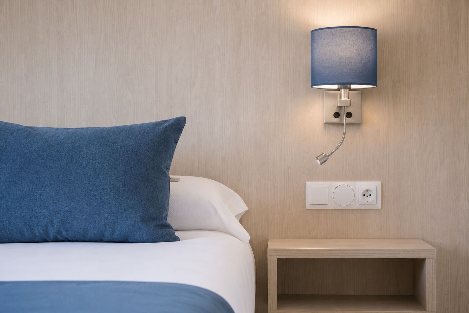 Una cama bien tendida con un almohá de color azul, una mesita de noche con una lámpara y un enchufe de pared.