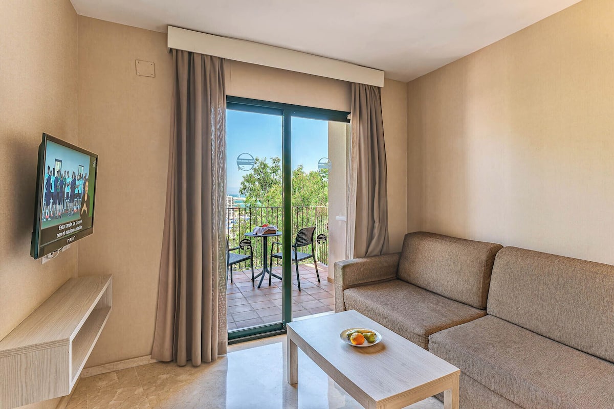 Une chambre d’hôtel moderne avec un canapé, une petite table sur laquelle sont disposés des fruits, un téléviseur fixé au mur et un balcon équipé d’une table et de chaises.