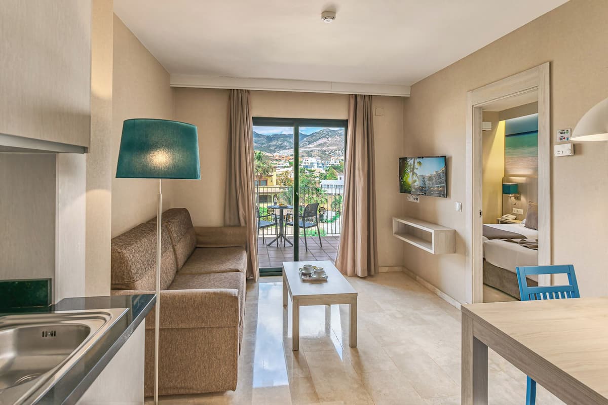 Une chambre d’hôtel équipée d’une kitchenette, d’un canapé, d’une table à manger, d’une télévision et d’un balcon offrant une vue sur la ville et les montagnes.