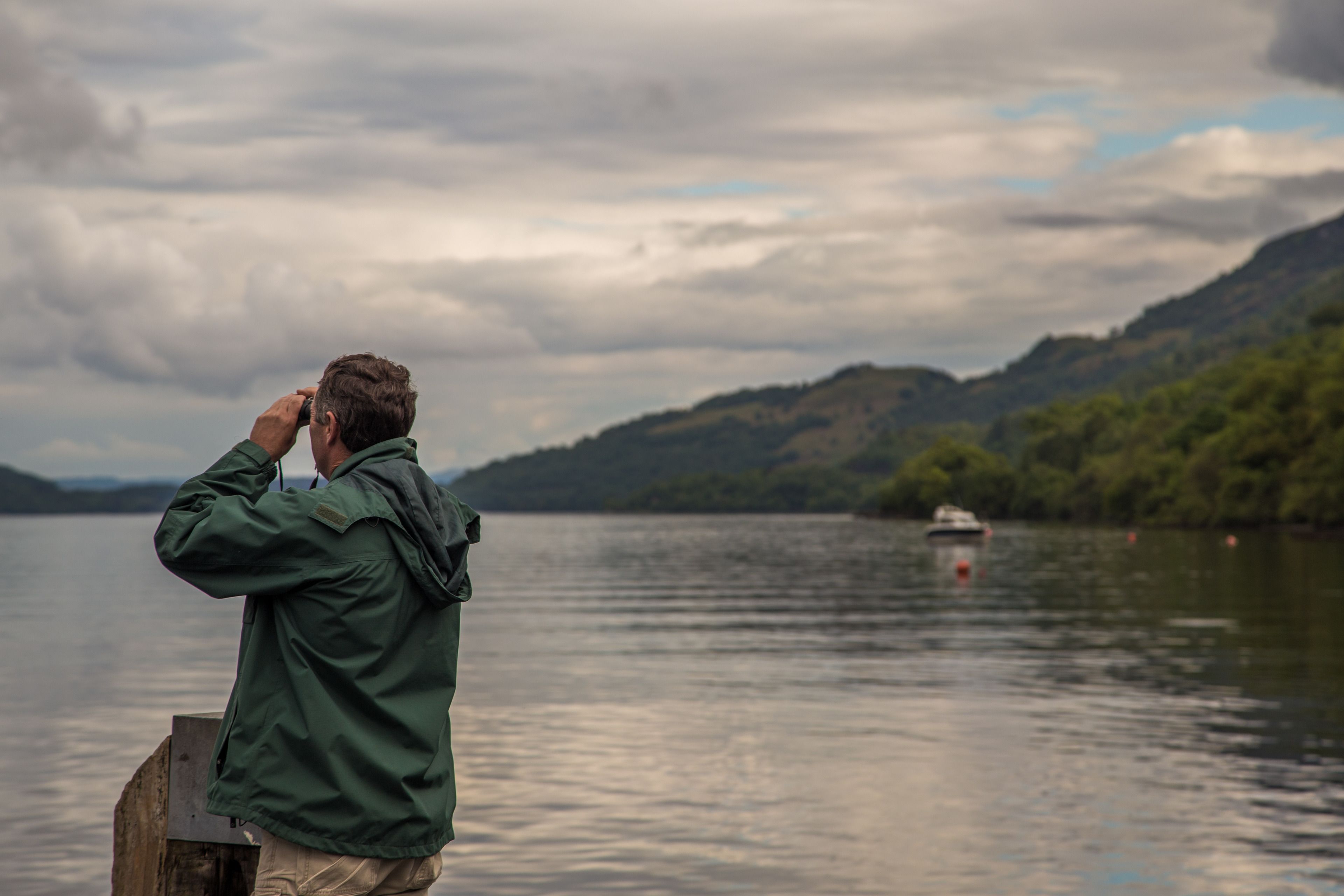 Una persona fotografiando un lago, con un barco a lo lejos y colinas al fondo.