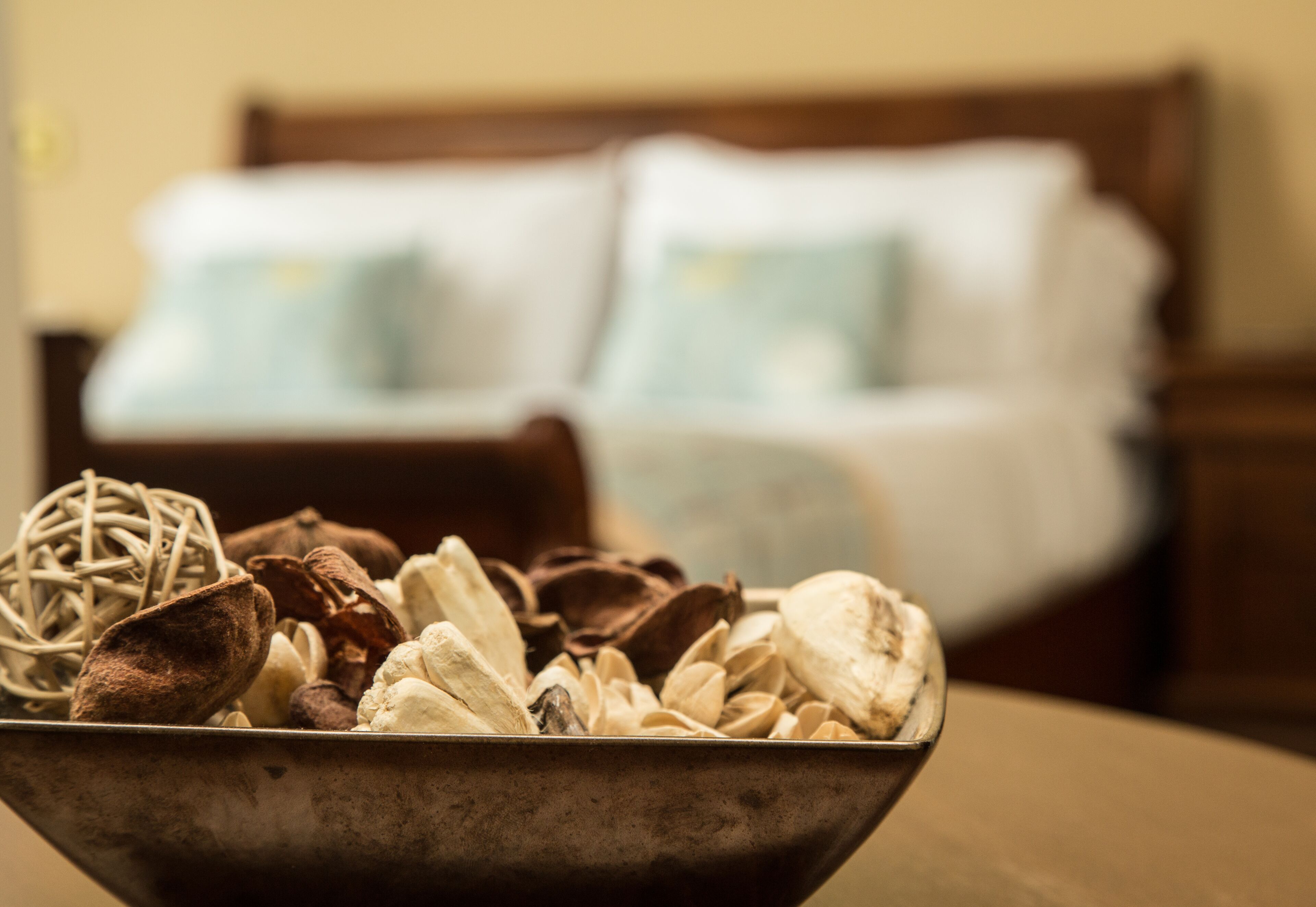 A metal bowl filled with various nuts and dried fruits on a table. In the background, there is a blurred view of a bed with pillows.