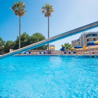 Una piscina con agua azul cristalina, rodeada de palmeras y un edificio con sombrillas azules y amarillas.