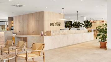 Un área de recepción con mostrador de madera, zona de estar con sillas y mesas, y una planta en maceta.