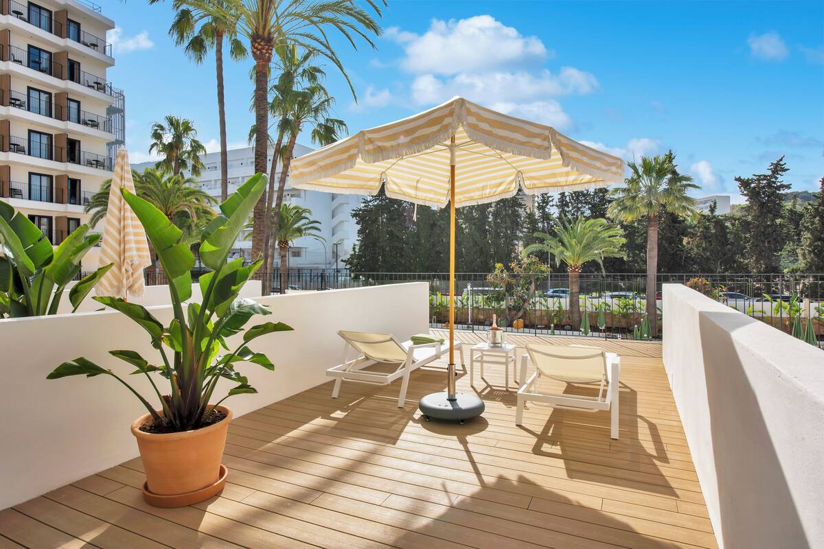 A balcony with a wooden floor, a potted plant, two white lounge chairs, a striped umbrella, and a tall palm tree.