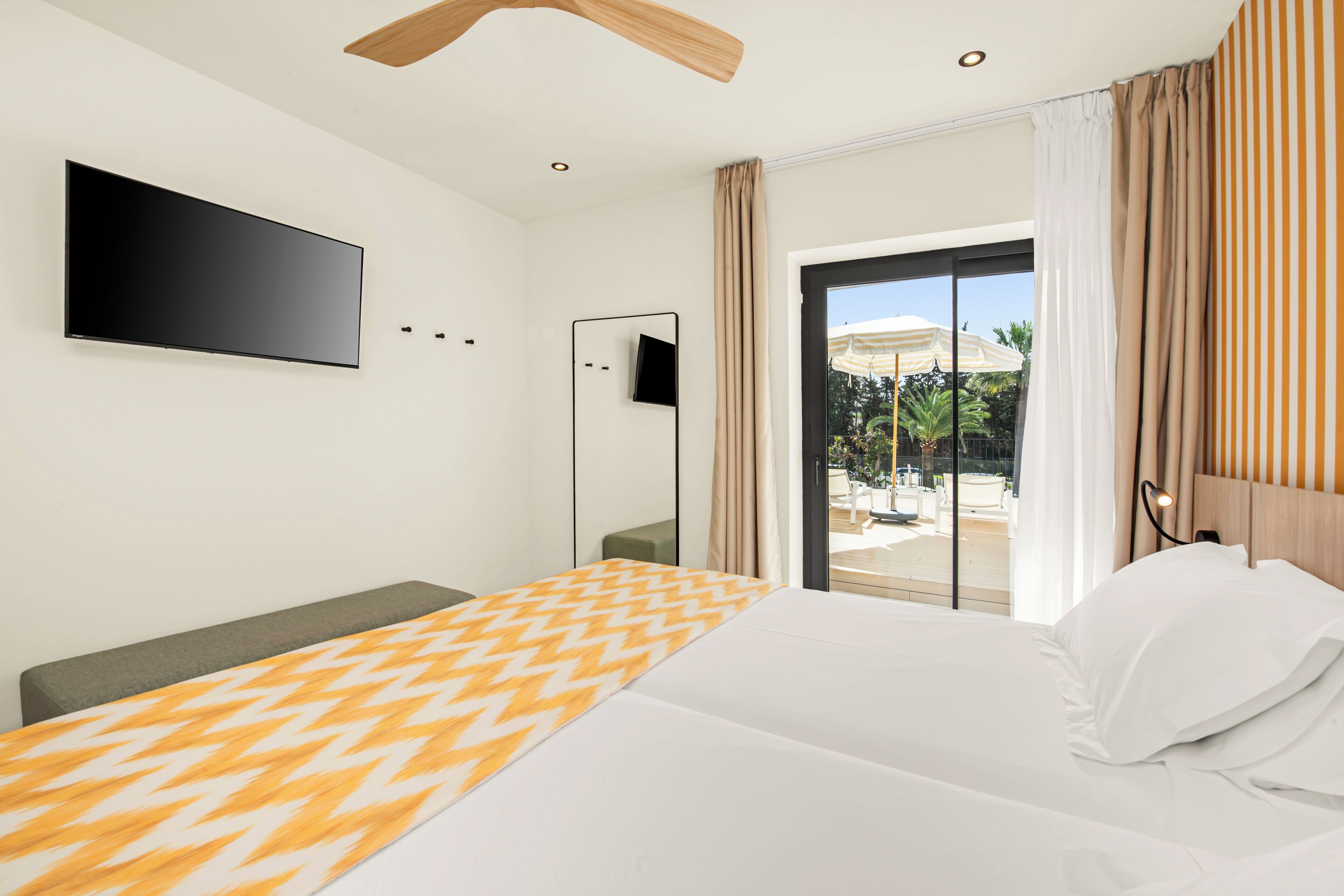A hotel room with a bed, a flat-screen TV, a ceiling fan, and a sliding glass door to a balcony.