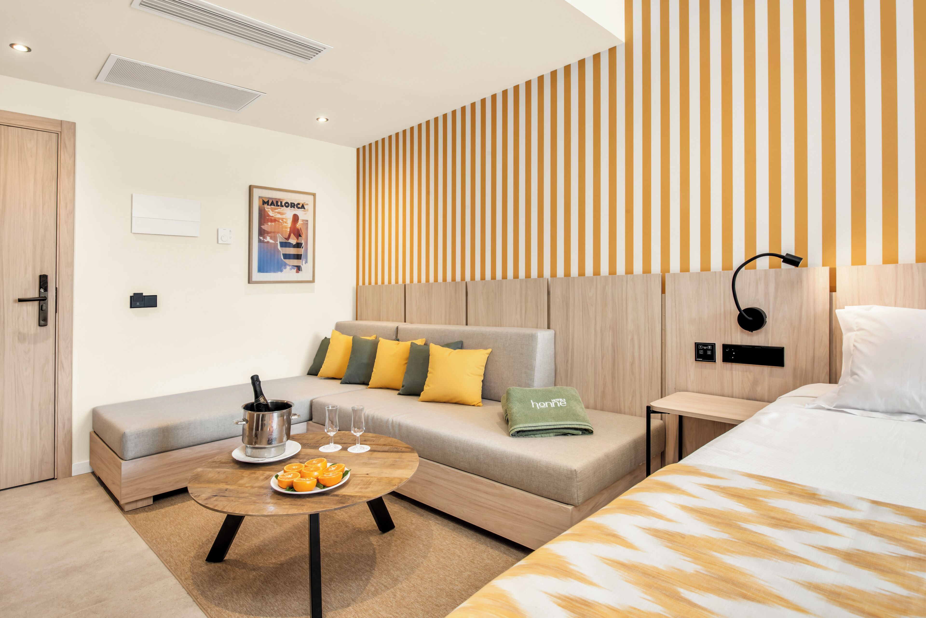 A modern hotel room with a sofa, a small table with fruit, a bed, and a wall-mounted lamp.