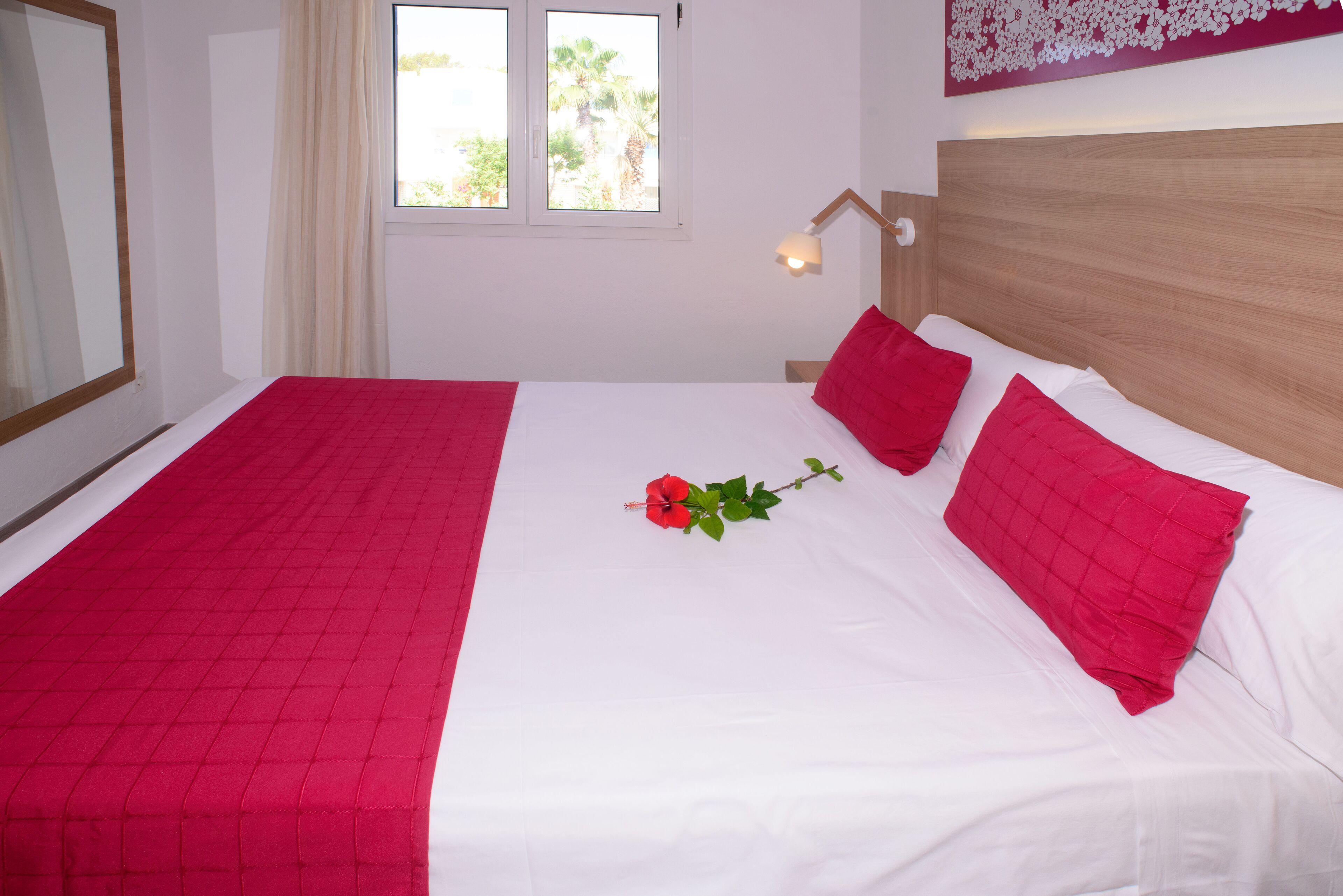 Una cama bien hecha con una colcha roja y blanca, una rosa roja solitaria y un cabecero de madera.