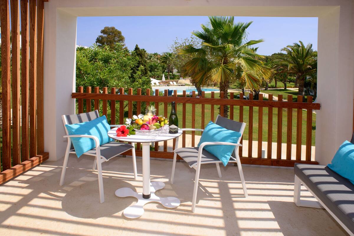 A balcony with two white chairs, a table with fruit and a bottle, and a view of a pool and palm trees.