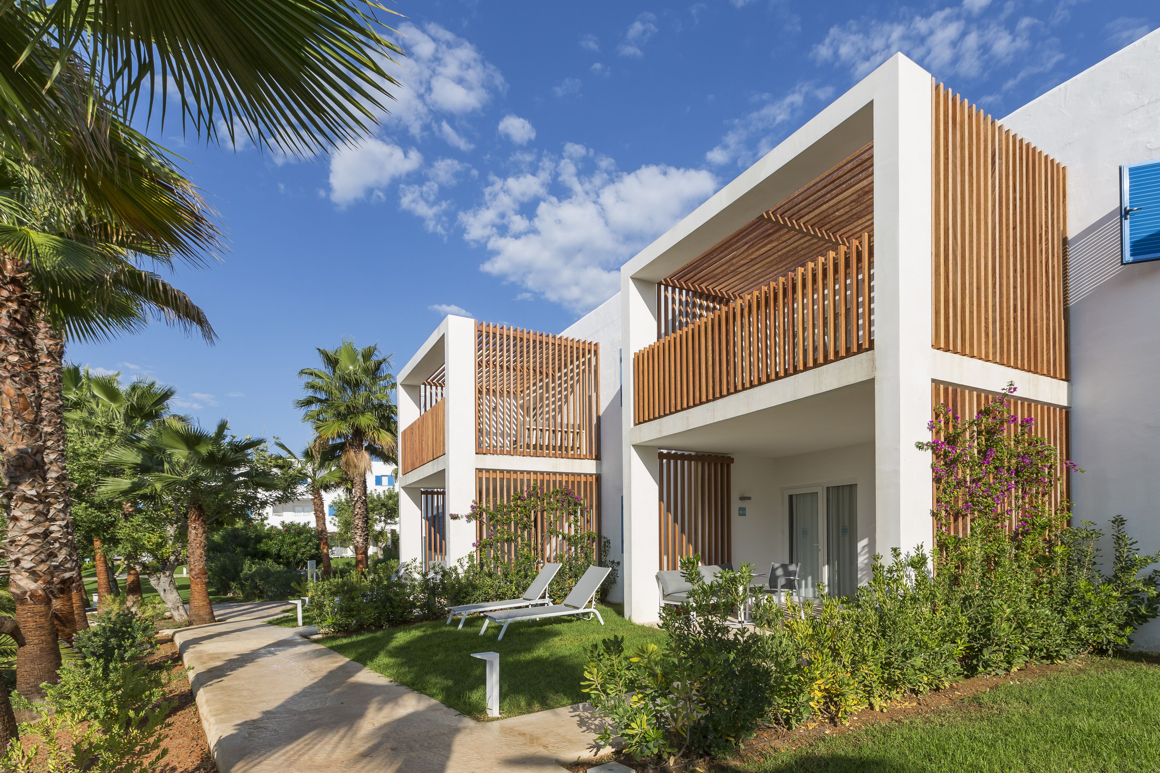 Un edificio moderno con fachada blanca, balcones con listones de madera y un jardín bien cuidado con palmeras y un sendero que lleva a sillones de descanso.