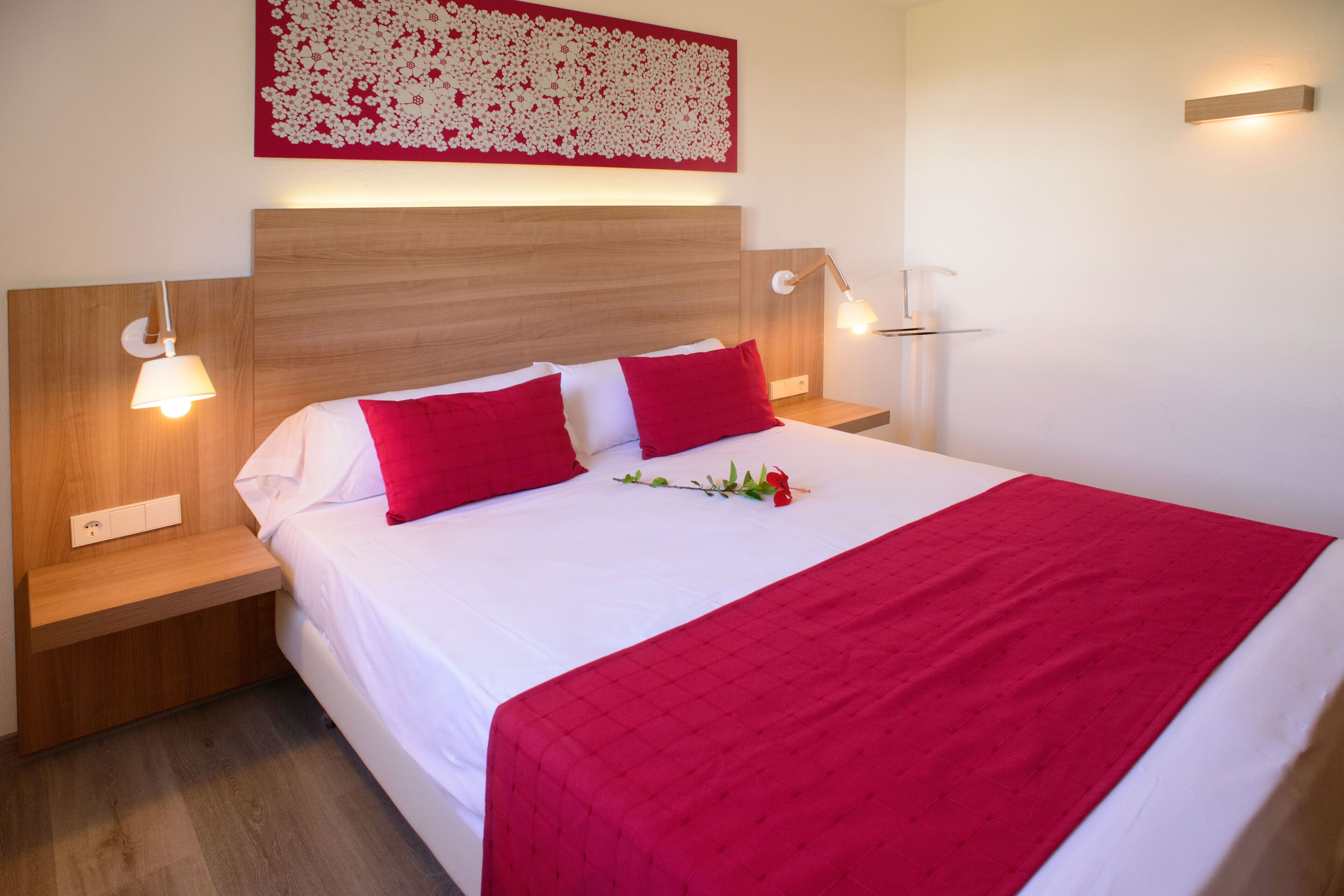 A neatly made bed with red pillows and a red blanket, wooden headboard, bedside lamps, and a floral artwork on the wall.