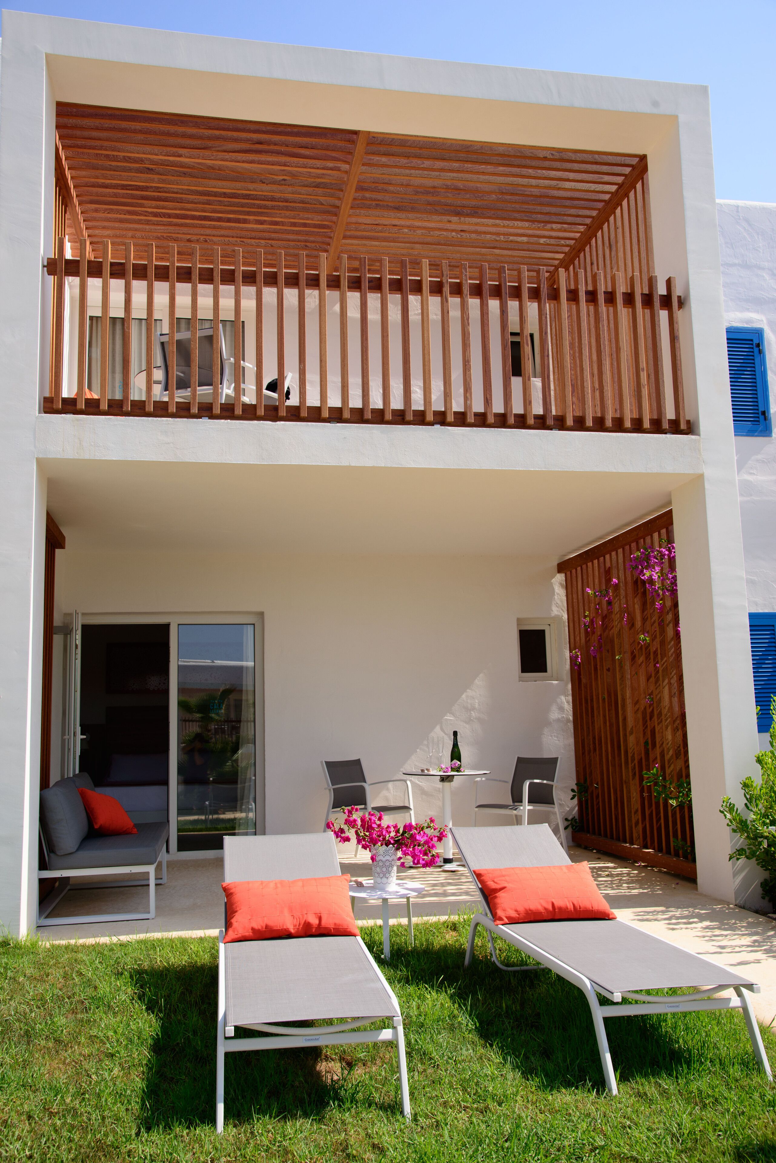 Una casa moderna de dos pisos con un balcón, una zona de estar con dos tumbonas, una mesa con sillas y un pequeño jardín.