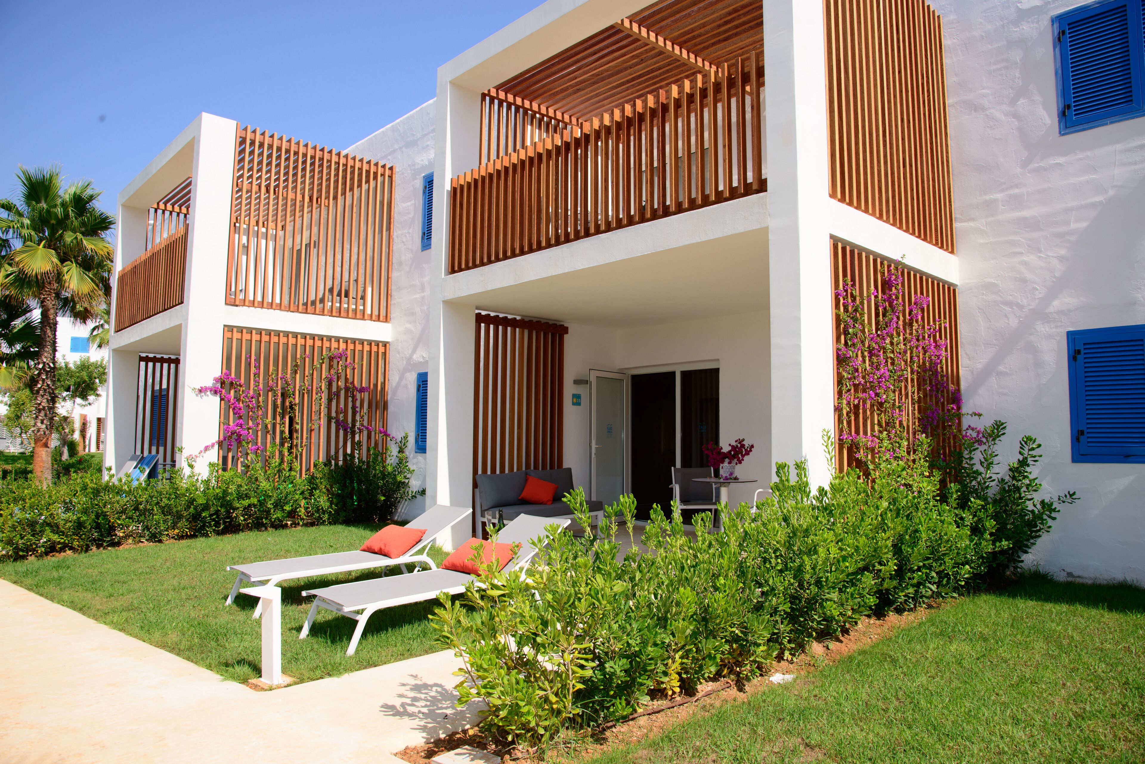 A modern two-story building with a balcony, outdoor seating area, and lush greenery.