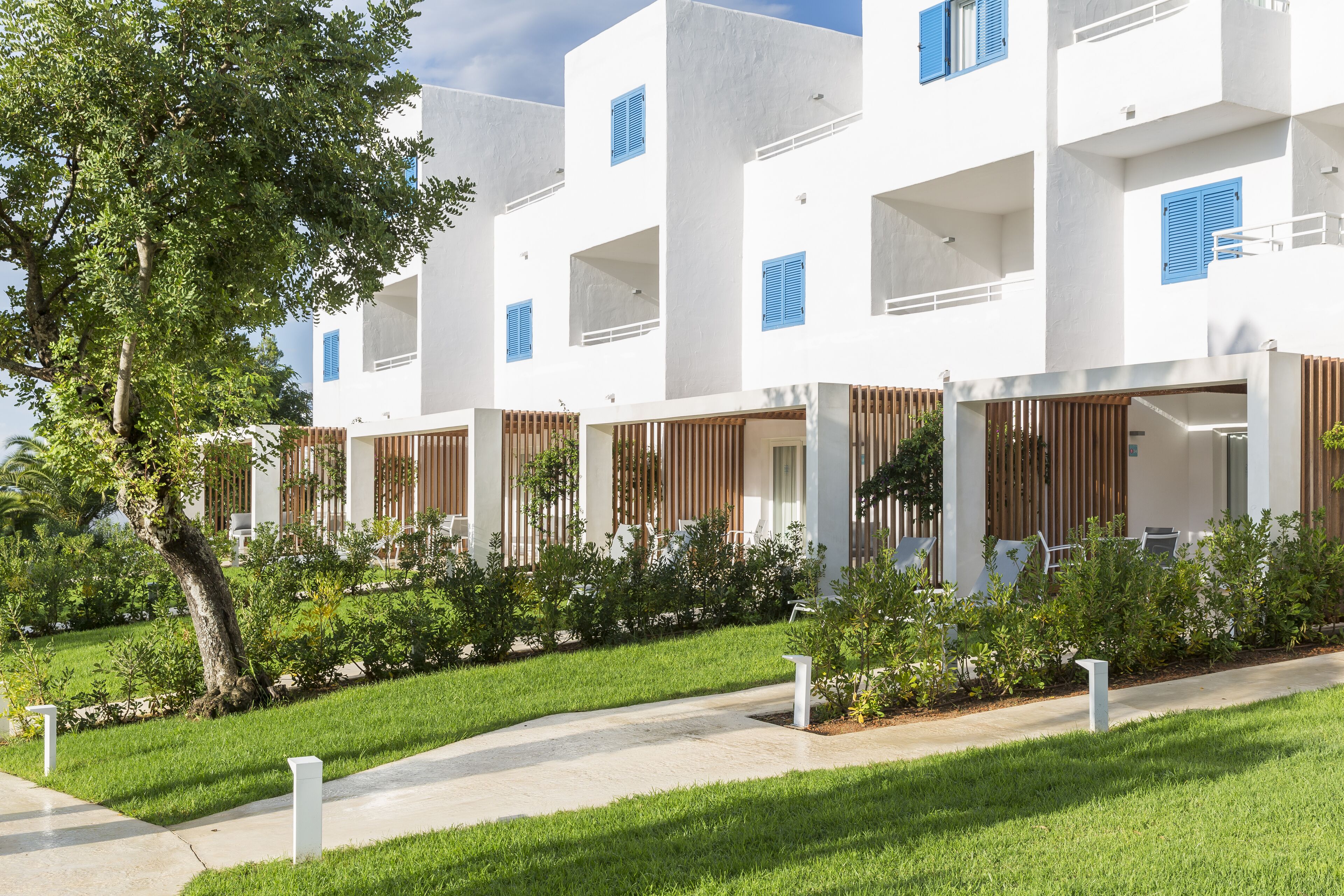 A modern building with white exterior walls and blue shutters, surrounded by well-maintained greenery and a paved pathway.