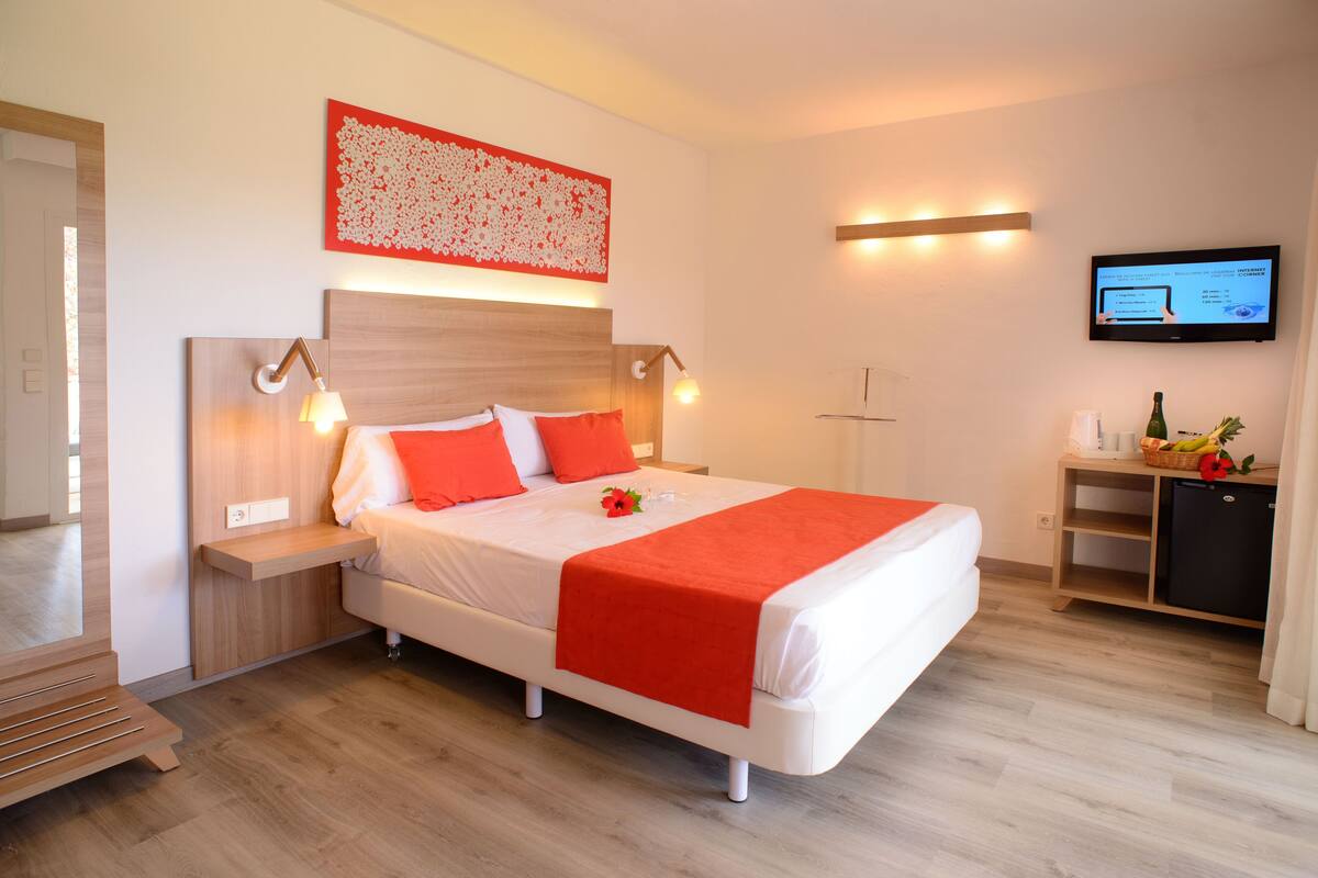 A modern hotel room with a large bed, a wooden headboard, bedside tables with lamps, a flat-screen TV, and a small refrigerator.