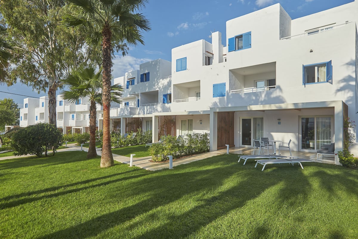 A modern building with white walls and blue shutters, featuring balconies and a well-maintained lawn with lounge chairs.