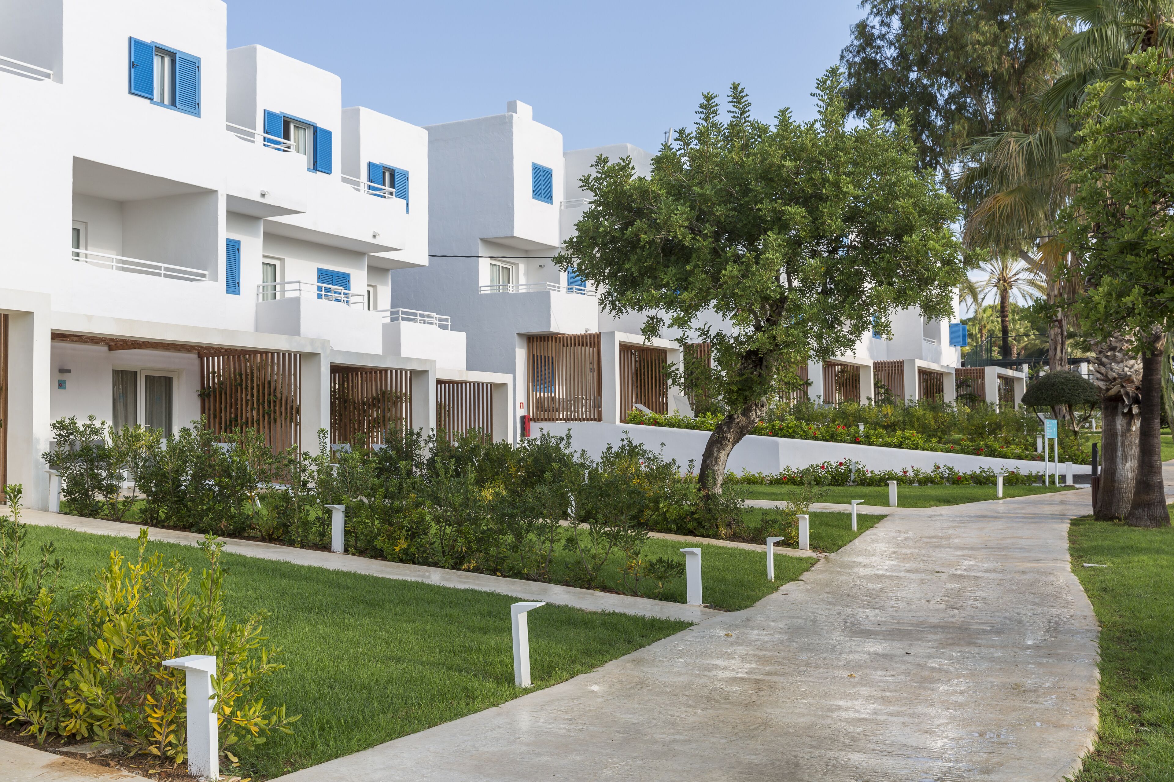 A row of white buildings with blue shutters, a concrete pathway, and green landscaping.