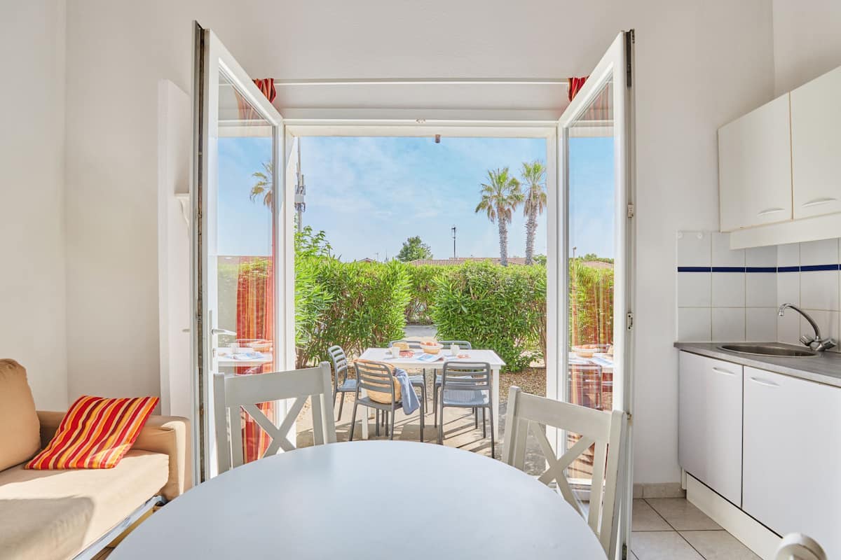 An open sliding door leading to a patio with a table and chairs, a kitchen area with white cabinets, and a view of greenery and a building in the distance.
