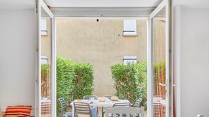 Eine offene Terrasse mit Tisch und Stühlen, ein Bett mit gestreiftem Kissen und Blick auf ein Gebäude mit Fenstern.