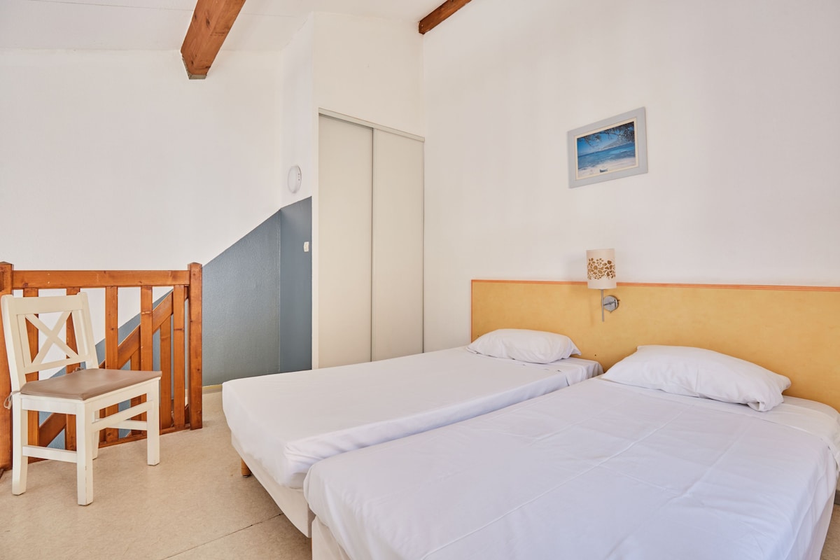 A room with two single beds, a wooden chair, a small table, and a picture on the wall.