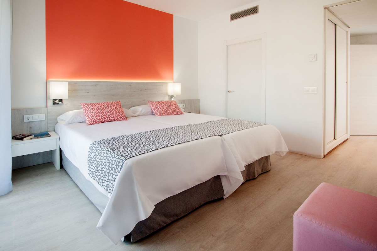 Une chambre d’hôtel avec un grand lit, deux tables de chevet, une commode, une chaise rose et une porte.