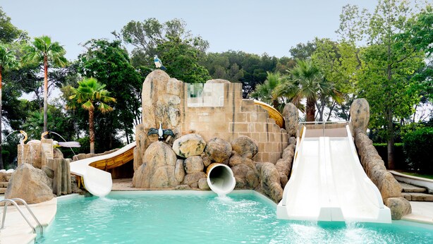 A pool area with a water slide, a rock wall, and palm trees. - Globales Hotel Maioris (Llucmajor)