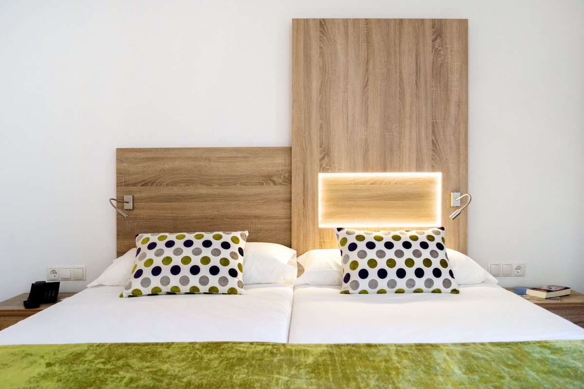 Cama de casal com roupa de cama branca, almofadas de bolinhas, cabeceira de madeira, abajures e manta verde.