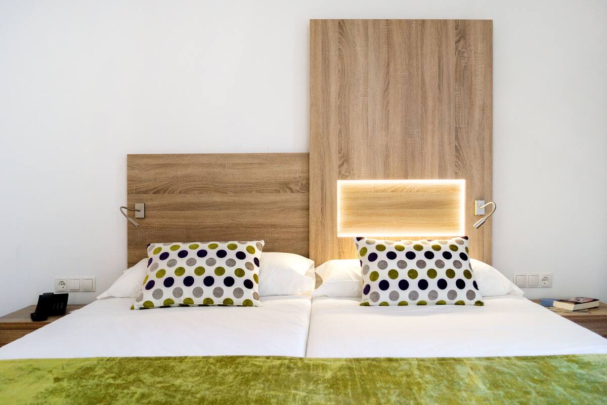 Une chambre d’hôtel avec une tête de lit en bois, deux lits blancs avec des oreillers à pois, un plaid vert et une table de chevet sur laquelle se trouvent un téléphone et un livre.