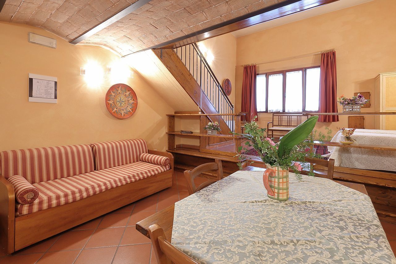 A cozy living room with a wooden sofa, a dining table with a patterned tablecloth, a vase with flowers, and a staircase leading to another level.