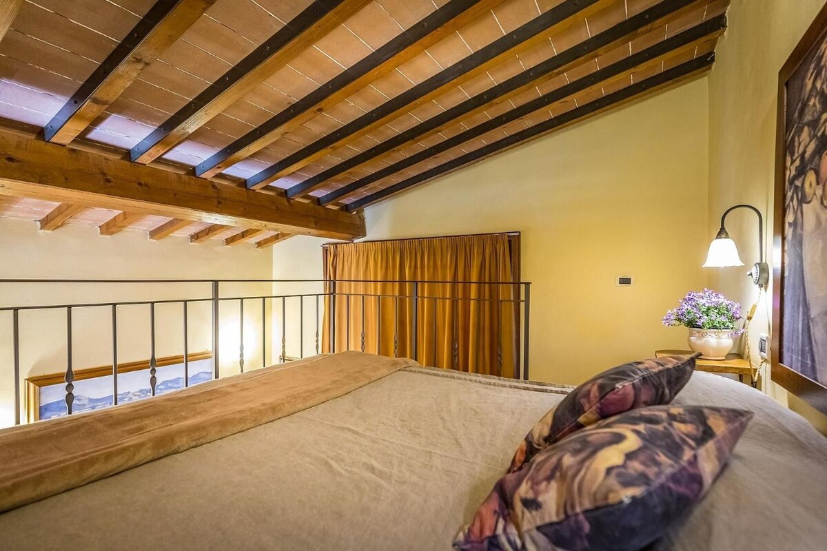 A bedroom with a wooden ceiling, a bed with a patterned pillow, a nightstand with a lamp, and a window with curtains.