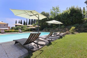 Un área junto a la piscina con sillones de descanso y sombrillas, rodeada de vegetación y un cielo despejado.