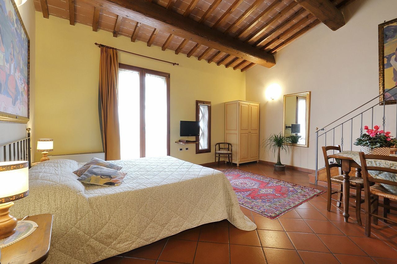 A bedroom with a bed, wooden ceiling beams, a window with curtains, a television, a cabinet, a chair, a table with a plant, and a rug.