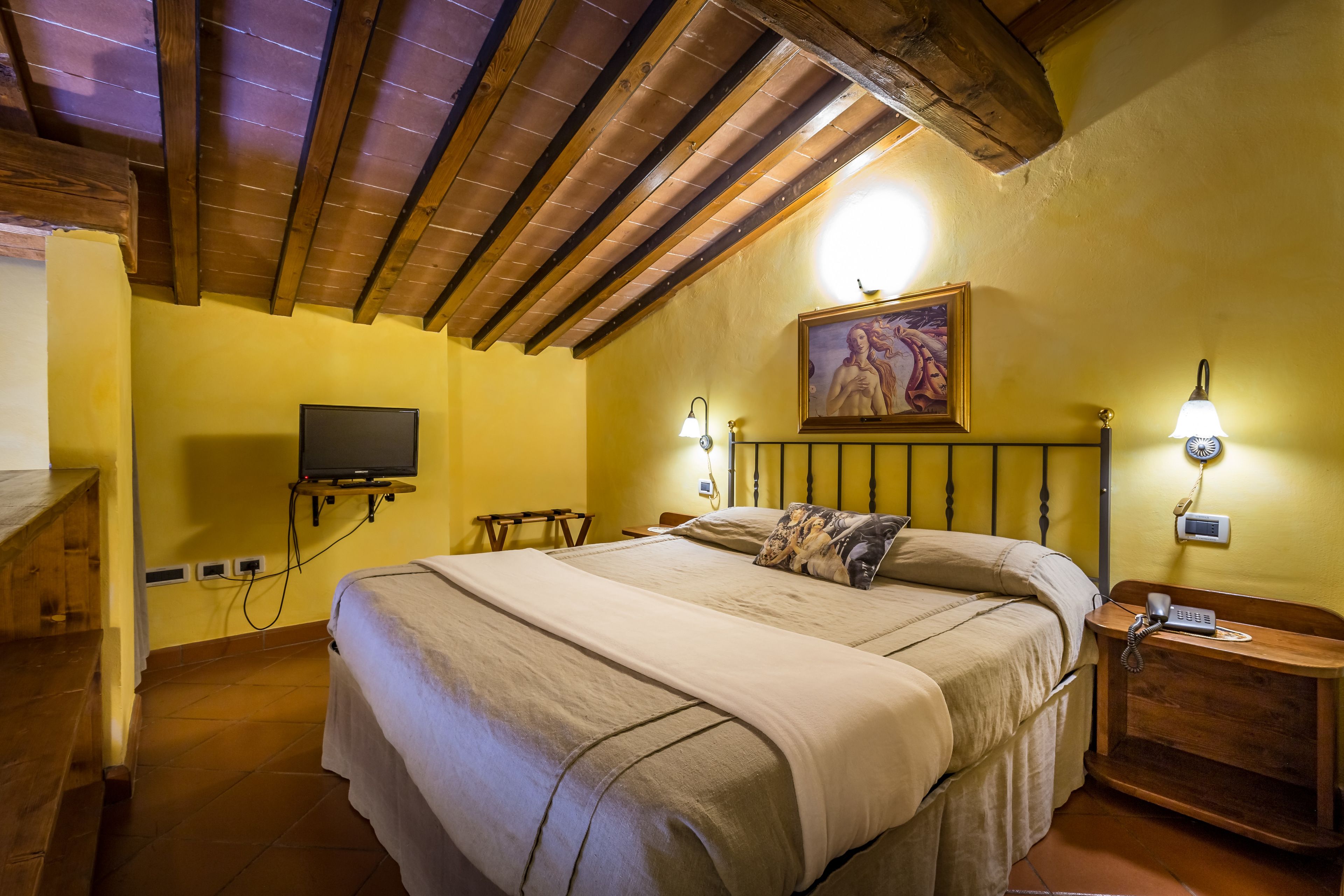 Une chambre avec un plafond en bois, un lit recouvert d’une couvre-lit beige, un tableau au mur et une table de chevet avec une lampe.