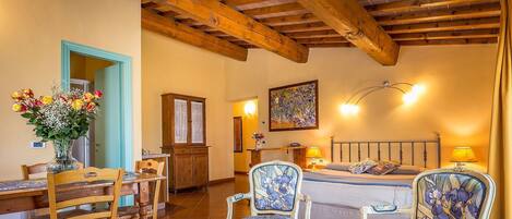 A cozy room with a wooden ceiling, a bed with a headboard, two upholstered chairs, a dining table with chairs, a cabinet, and a painting on the wall.