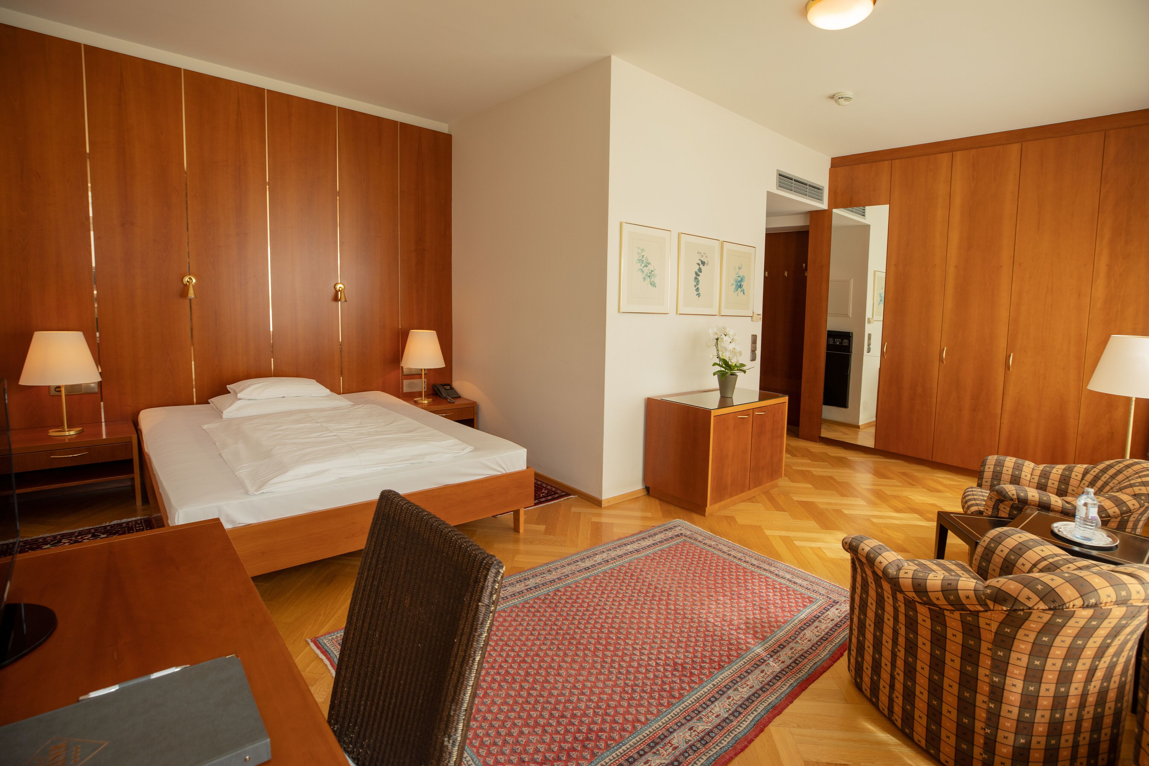 A hotel room with a large bed, wooden wardrobe, a desk with a chair, a small table, and a sofa.