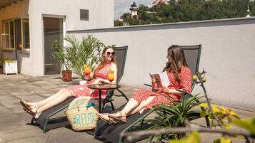 Two people relaxing on sun loungers in an outdoor seating area with potted plants and a view of a hill with buildings.